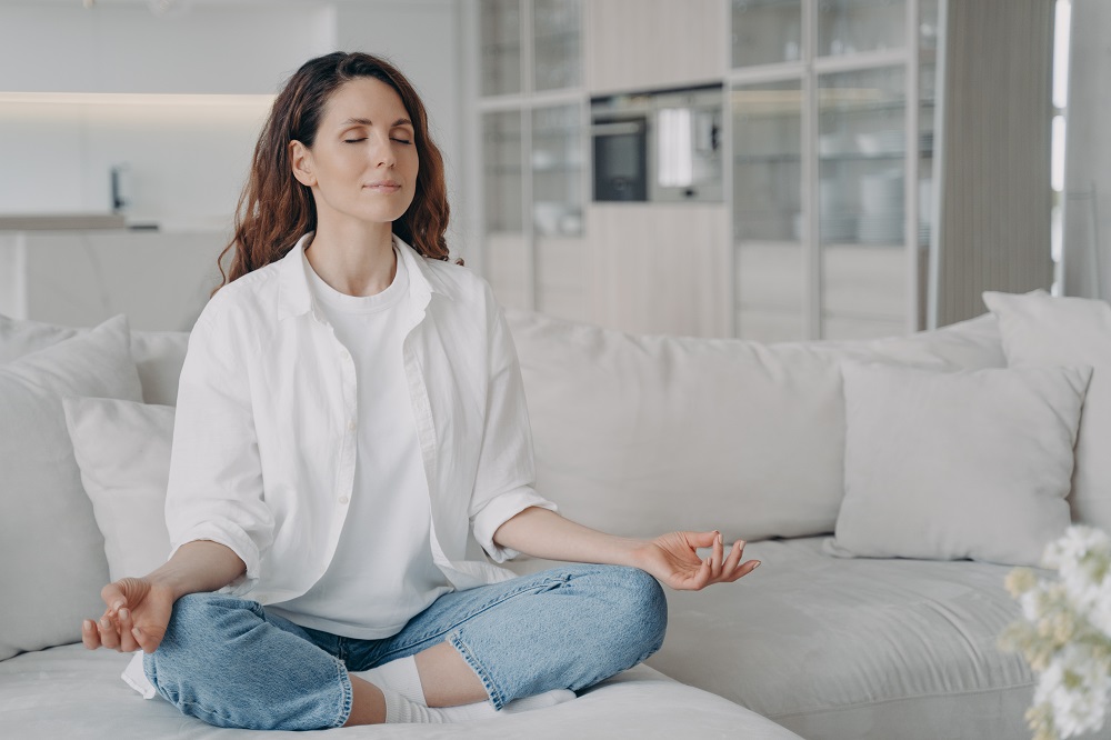 Mulher pratica ioga sentada no sofá em pose de lótus. Hábitos saudáveis, estilo de vida consciente, alívio da ansiedade
